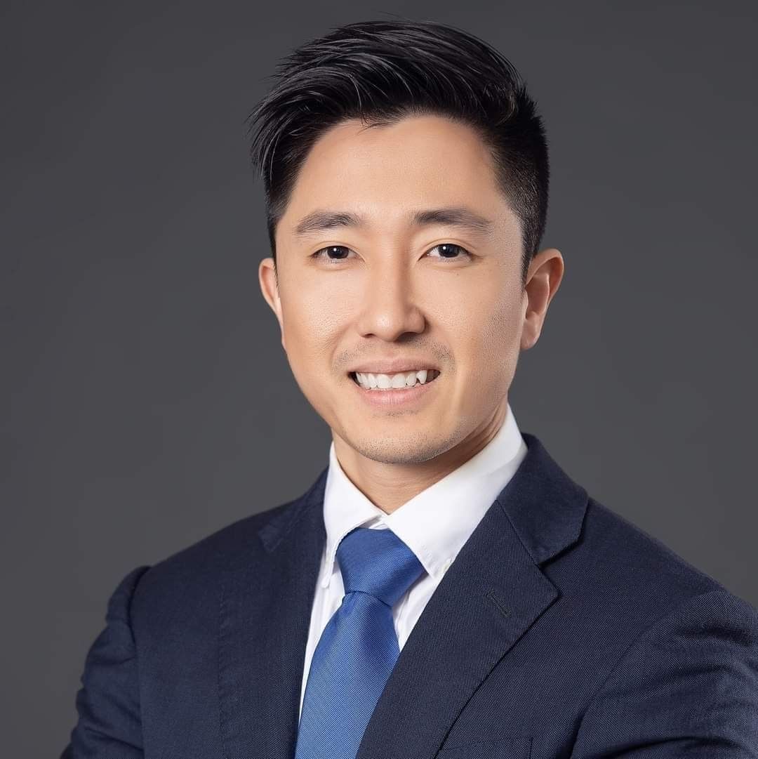 Smiling man in a dark suit and blue tie posing with a gray background.