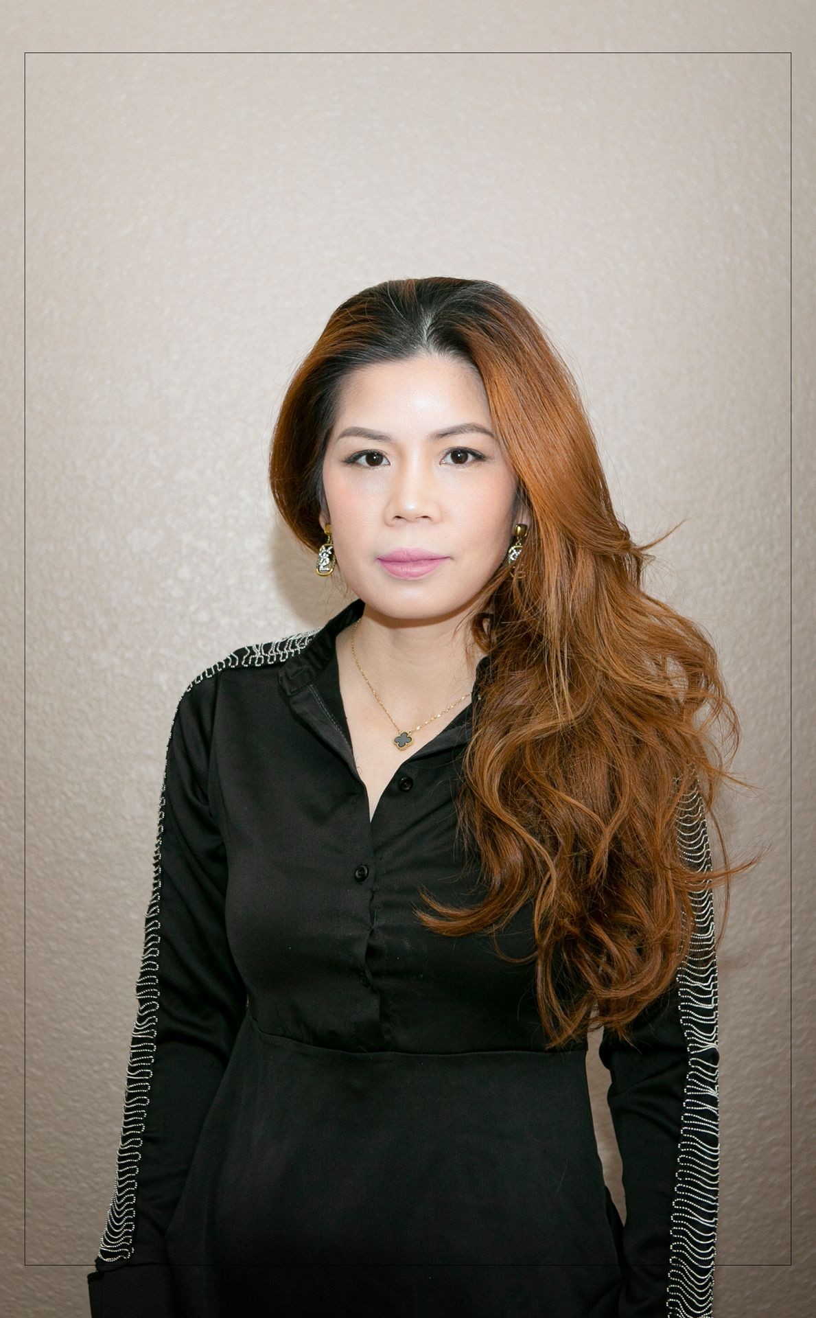 Person with long hair wearing a black top, standing against a beige background.